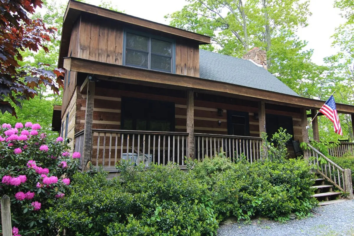 The front exterior of a log cabin with a welcoming porch, an American flag, vibrant flowers, and cozy seating—inviting residents and guests into a friendly neighborhood atmosphere.