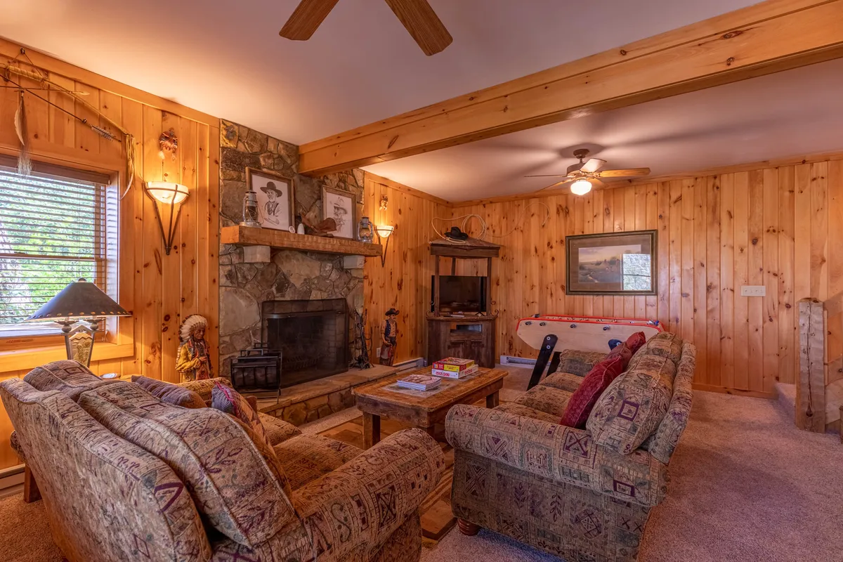 A welcoming great room featuring a prominent stone fireplace framed by rustic wooden beams, with a cozy seating area arranged around the hearth. The wooden furniture and natural textures create an inviting atmosphere, ideal for gatherings, relaxing, and enjoying the mountain ambiance.