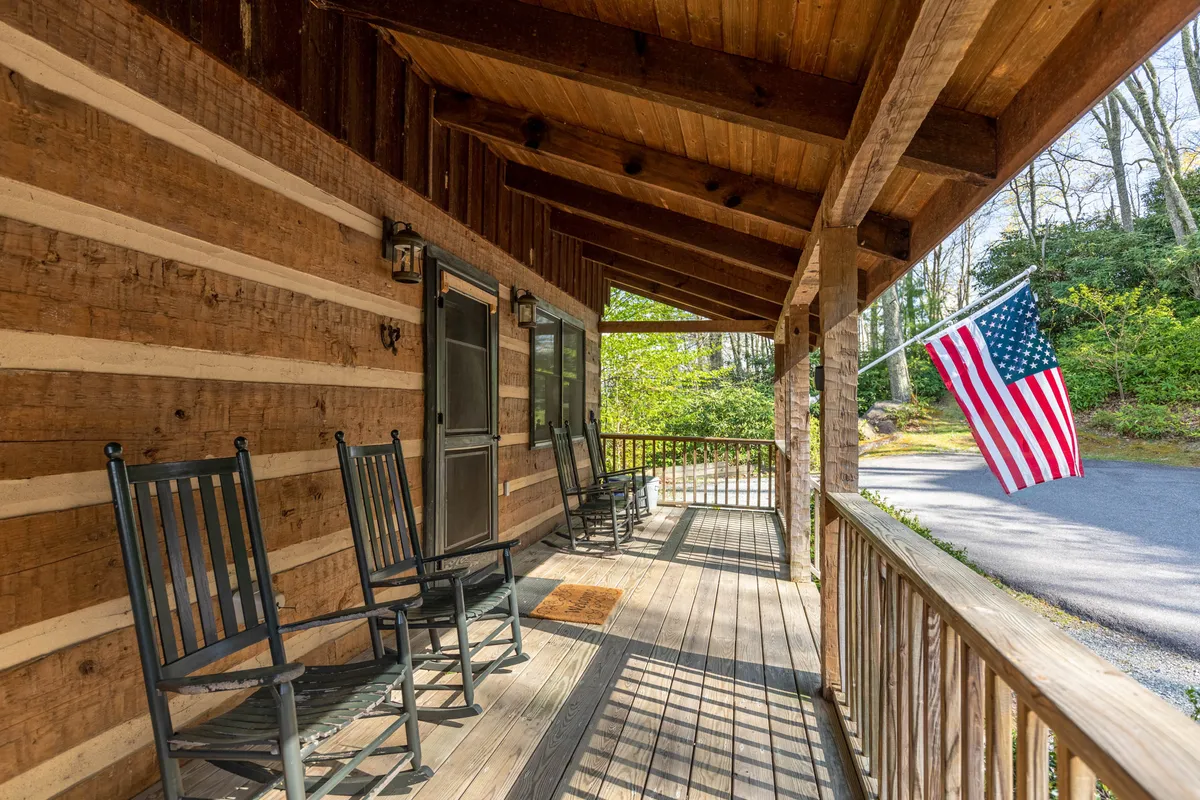 The image captures a peaceful porch scene featuring rocking chairs lined up beneath a wooden structure, with an American flag gently waving. The vantage invites guests to imagine quiet moments watching the sunset or listening to the sounds of nature.