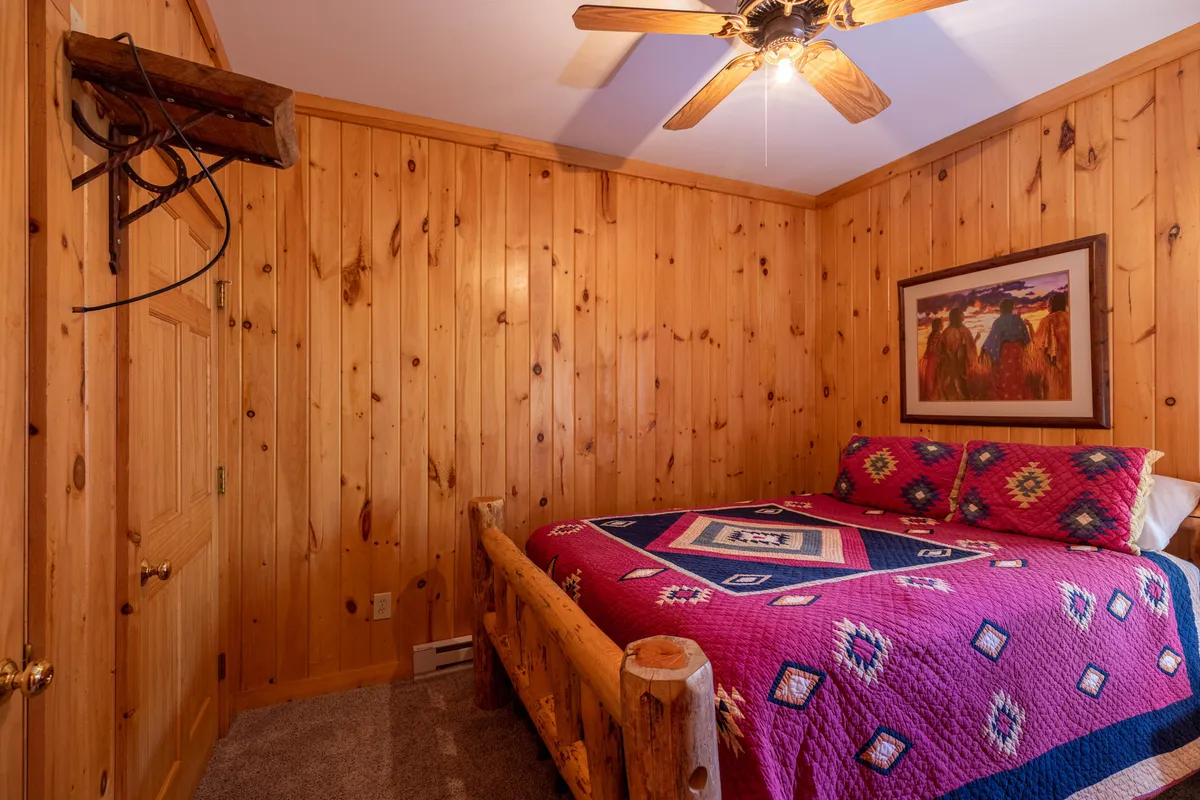 A cozy bedroom featuring a rustic wooden bed frame, a vibrant colorful quilt, and warm wood paneling. Framed artwork hangs on the walls, and soft natural light filters through a window, creating an inviting and restful atmosphere unique to a master suite.