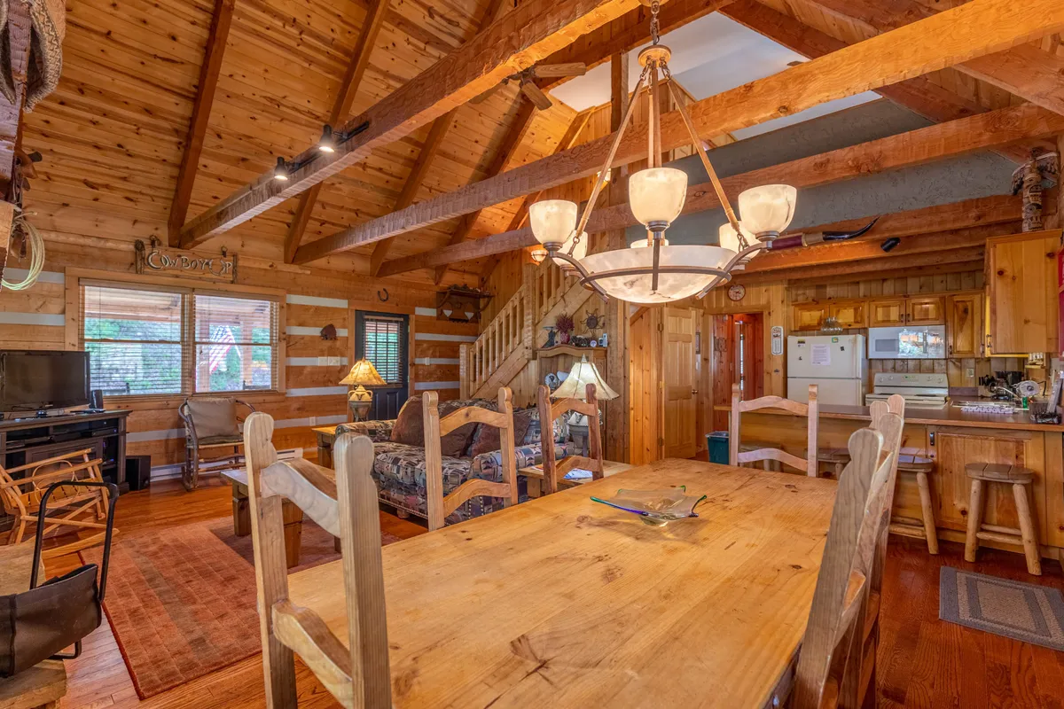 A large wooden dining table beneath a chandelier sits adjacent to a sofa and a fireplace, with rustic elements throughout. The space is designed for communal activities like shared meals, late-night stories, and group games, evoking a sense of togetherness and warmth.