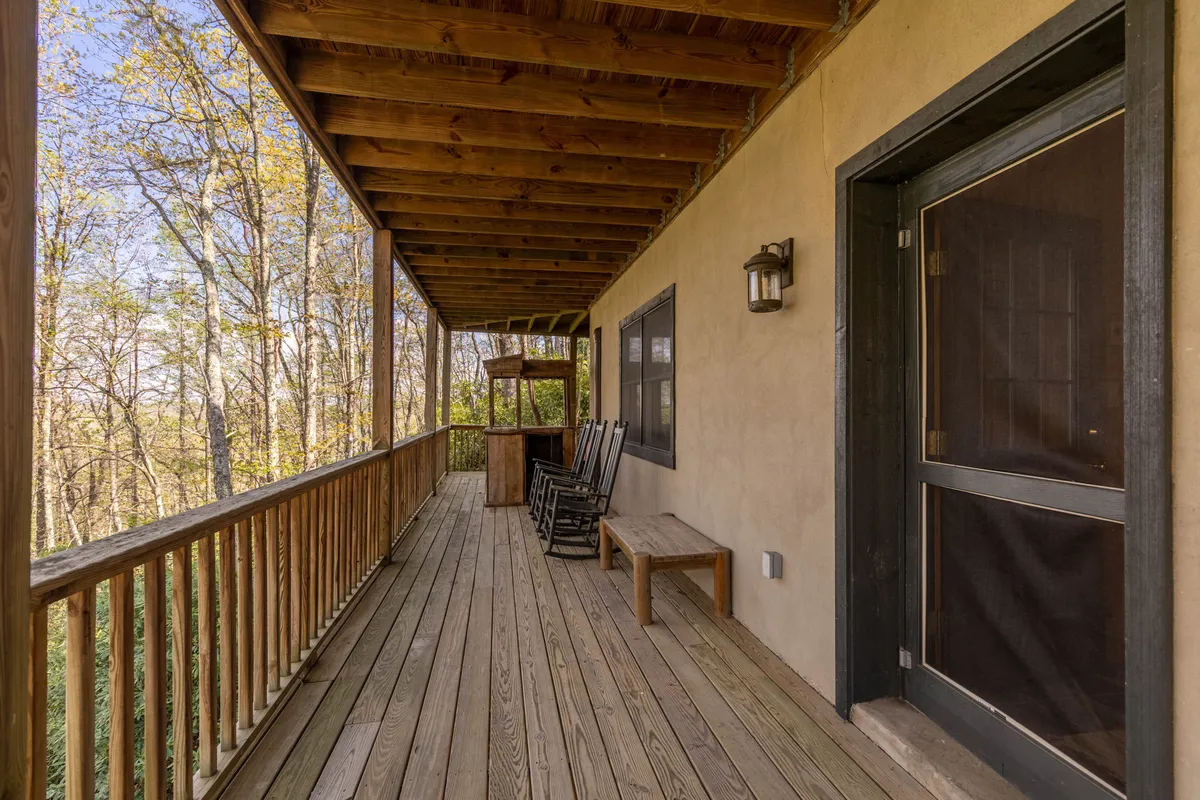 A serene exterior view of a wooden deck furnished with classic rocking chairs, a wooden railing, and surrounded by lush trees. The area includes a small bar, suggesting both relaxation and opportunities for socializing outdoors.
