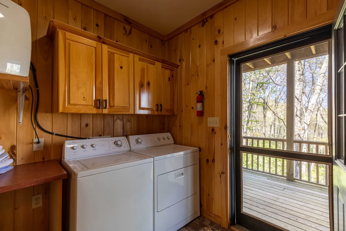 A practical laundry area featuring a washing machine, dryer, and wooden cabinetry, with a screen door that leads outside and hints at tranquil nature views beyond. The presence of the door creates a subtle connection between interior comfort and outdoor exploration.