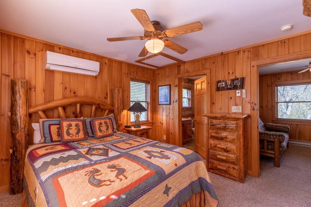 A bright bedroom with a log bed frame, colorful quilt, wooden bedside furniture, and a table lamp. Sunlight streams in, highlighting the inviting textures and warm, rustic design elements that echo the retreat's natural setting.