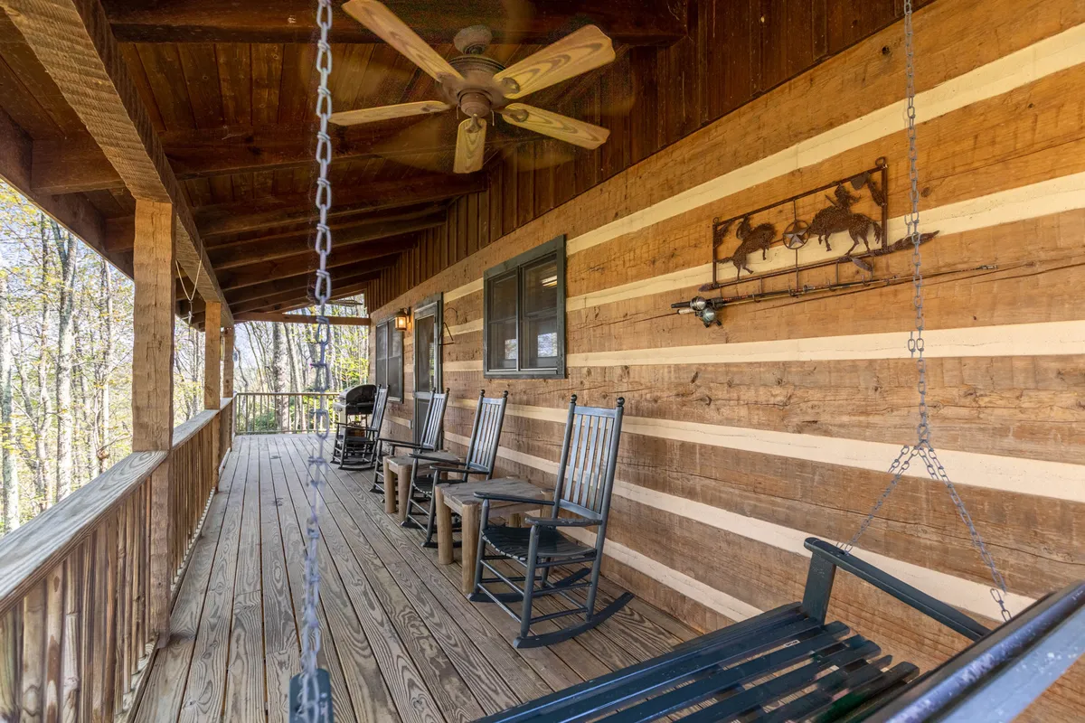 This exterior image features a wooden deck adorned with classic rocking chairs, a ceiling fan overhead, and decorative wall art, offering views out to the surrounding natural scenery. The warm wood tones and comfortable seating invite guests to unwind while taking in the fresh mountain air.