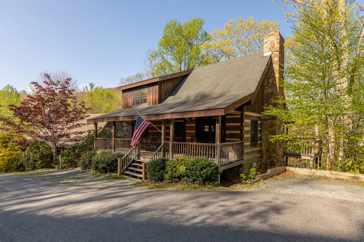 The image showcases a classic log cabin nestled in the North Carolina mountains, featuring a sturdy stone chimney, a welcoming front porch draped with an American flag, and a backdrop of natural beauty ideal for gatherings and relaxation.