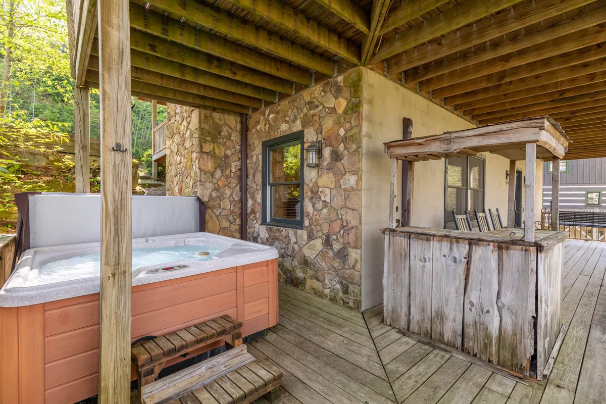 An inviting outdoor space featuring a hot tub bordered by a natural stone wall, a wooden bar area for drinks, and views of surrounding foliage. The setup offers a tranquil retreat after outdoor adventures.